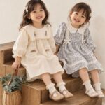 Two smiling toddler girls sitting on wooden steps wearing floral cotton dresses and cute shoes.
